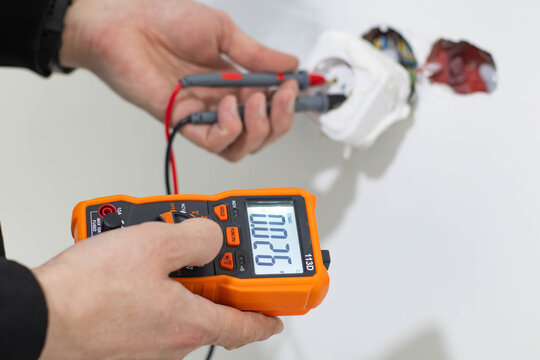 Close-up of a professional technician's hands using a digital multimeter to check the voltage in a power socket during home electrical repair or maintenance. 