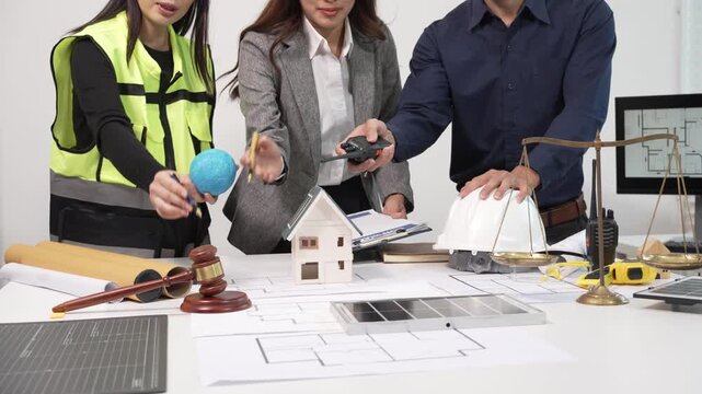 Architects and engineers collaborating at desk with blueprints, house model, hardhat, gavel and scale, discussing construction plans and costs in office meeting.