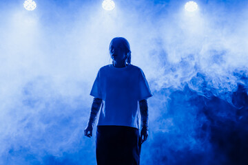 Performer stands under bright stage lights surrounded by dense blue smoke, creating a dramatic concert or theater atmosphere © ugis