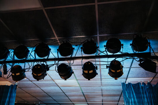 Black stage lights and cables mounted on an overhead rig, with blue backlighting creating a dramatic event production atmosphere