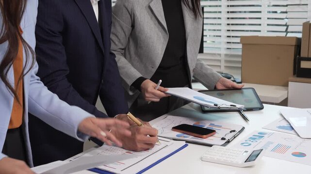 Business team collaborating at office desk reviewing financial charts, tablet and calculator, planning strategy in modern workspace with documents and reports.