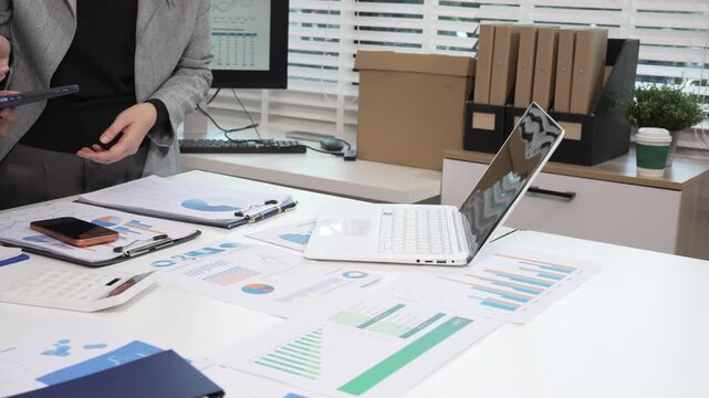 Business meeting at office desk with laptop, tablet, financial reports and charts, professional presenting data analysis and strategy planning.