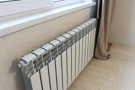 modern white bimetallic radiator mounted under a window, featuring sleek vertical fins; positioned beside a beige curtain, on light wood flooring, against textured walls