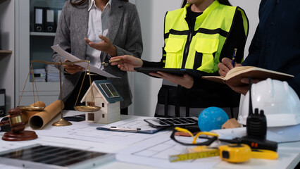Naklejka premium Team meeting with engineer in safety vest reviewing plans at desk with scale model house, hard hat, tools and documents; architecture, construction and project management concept.