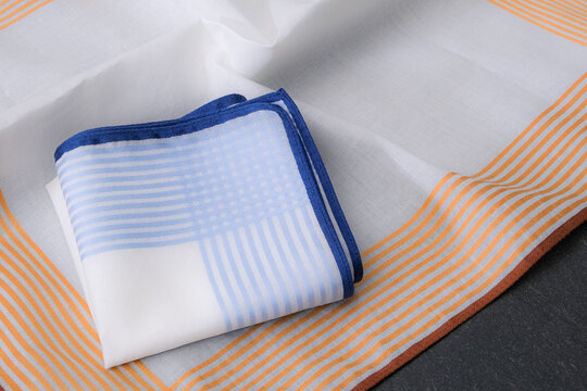 Close-up folded white handkerchief with blue stripes and hand-rolled edge on unfolded orange striped border handkerchief.
