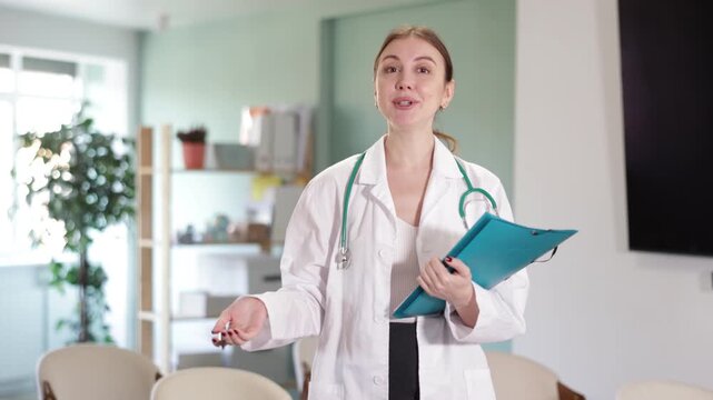 Portrait of young coach female doctor standing with folder with documents and materials in hands and talking in corporate seminar and inviting in light auditorium