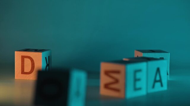 Hands rearrange blocks with letters to form the word dream in a colorful and artistic way. The activity involves focusing on letters, patterns, and movements in a studio-like space