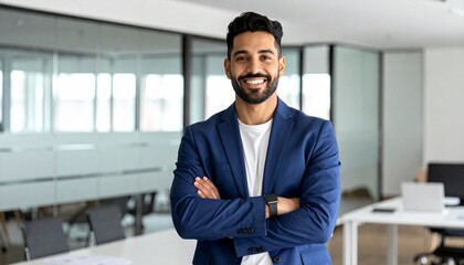 Fototapeta premium A smiling man in a blue blazer stands with arms crossed, in a modern, glass-walled office