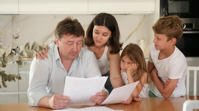Upset father reads and looks through financial documents. Sad wife with children in the background. High quality 4k footage