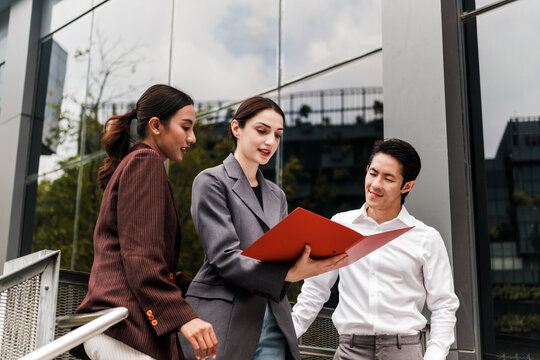 Three professionals outside a modern glass office review a red folder on the stairs, collaborating and discussing project plans.