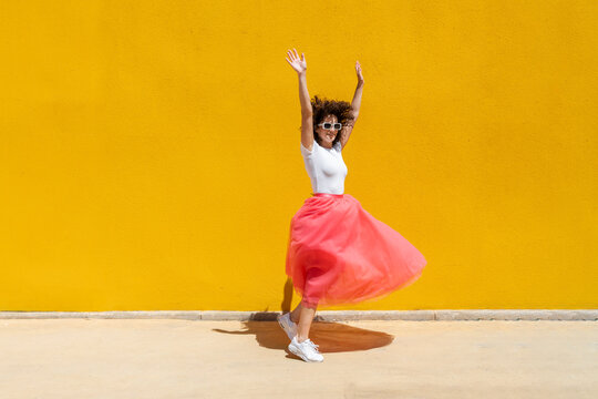 Cheerful woman dancing with arms raised near yellow wall