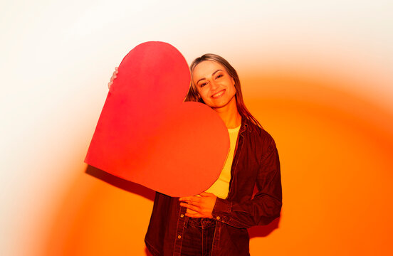 Smiling woman in jeans holding red paper heart indoors