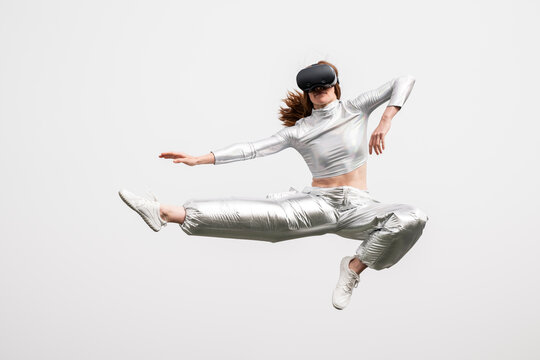 Redhead woman in metallic clothes jumping with VR headset outdoors