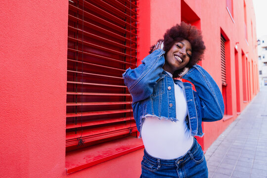 Smiling model wearing denim jacket listening to music by red wall