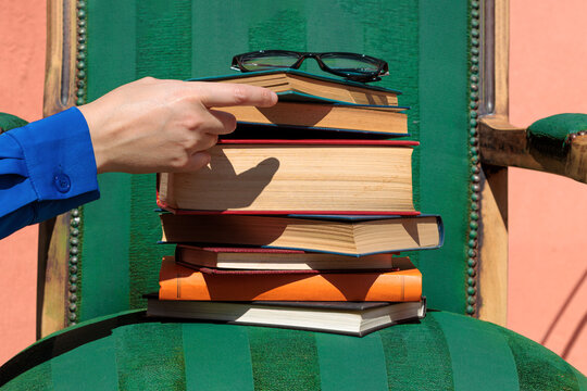 Hand touching stacked books on a green armchair with reading glasses