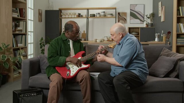Front view shot of adult Black man learning to play electric guitar while sitting on sofa at home male music teacher explaining technique Useful for home education services or tools