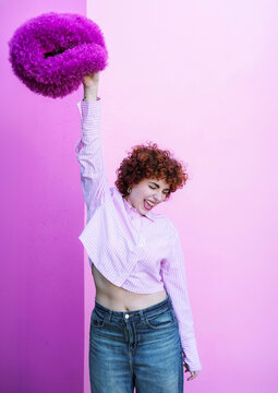 Redhead holding purple fluffy hat laughing against pink background
