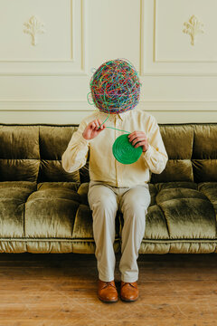 Man with mask of chaotic thoughts holding bundle of wires sitting on sofa