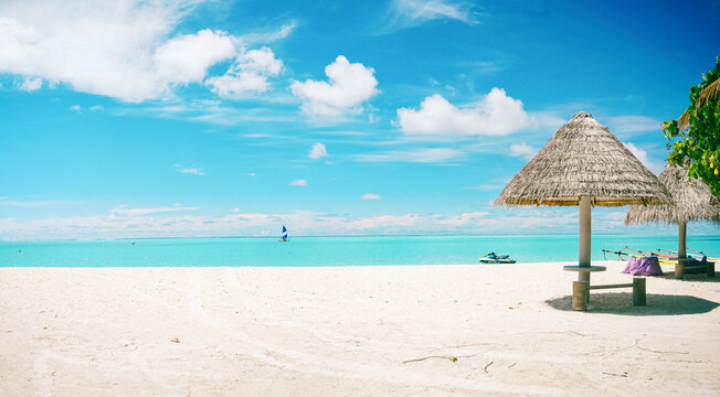 Panoramic nature view of a pristine Maldives beach featuring white sand and straw umbrellas on the shore. Turquoise ocean water under a bright blue sky with fluffy clouds.