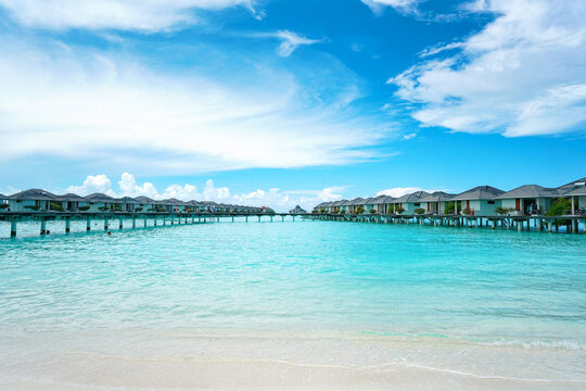 Panoramic nature view of luxury water bungalows stretching across the turquoise Maldives lagoon. A bright and serene scene with white sand in the foreground and a blue cloudy sky above.