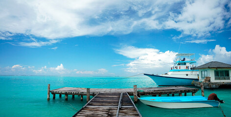 Panoramic nature view of a wooden pier with a small boat and a larger white yacht anchored in the crystal clear turquoise water of the Maldives. © Laura Pashkevich