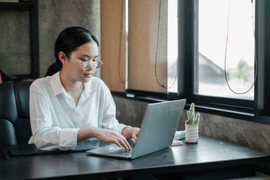 A focused woman in glasses works on her laptop in a contemporary office setting, surrounded by natural light and minimalist decor.