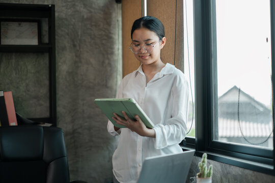 A young woman in a white shirt uses a tablet in a modern office setting with large windows and natural light.