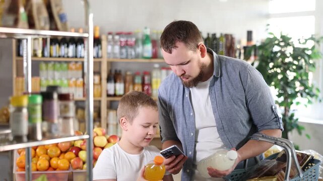 Positive young man with interested preteen son making purchases in local grocery store, using smartphone to scan barcodes on colorful bottles of sweet drinks for online payment