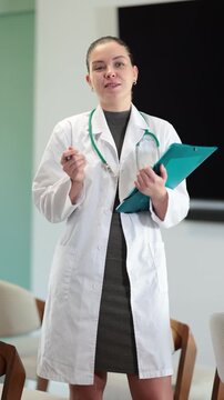 Portrait of young coach female doctor standing with folder with documents and materials in hands and talking in corporate seminar and inviting in light auditorium