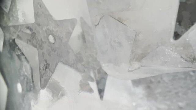Abstract macro video of silver star sequins moving between ice cubes with dreamy soft focus, ideal for winter, festive, and luxury visuals.