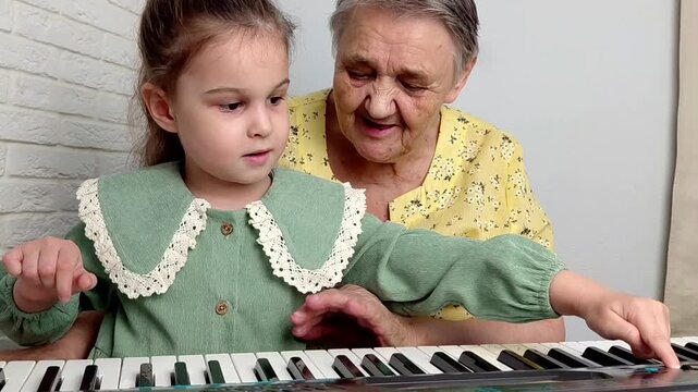 Elderly grandmother teaching little sweet girl playing piano, smiling with happiness, staying at cozy home, spending time together. Family, Love, Education Concept.