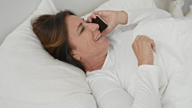 Woman lying in bed holding smartphone to ear with right hand, smiling under white duvet in a bedroom building; contentment connection.