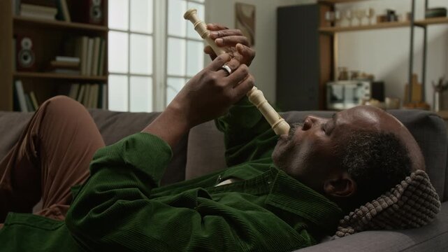 Side view shot of senior Black man playing recorder while lying on sofa at home enjoying music and relaxing, copy space Ideal for active seniors wellness and enrichment programs