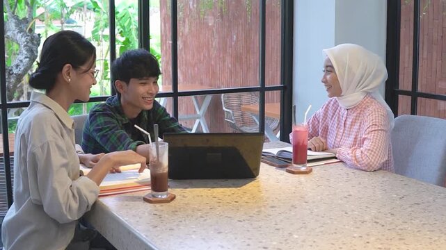 Three asian students study at cafe young hijab woman and two male peers exchange fist bump over laptop and notebooks, sipping