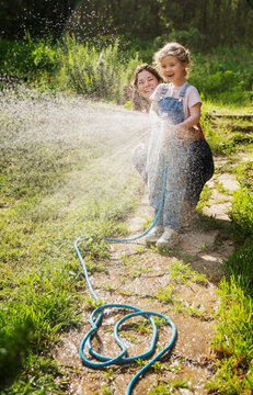 Happy mother and little daughter in denim overalls laughing and watering lawn with hose together.
