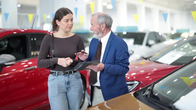 Elderly car salesman discusses terms and demonstrates car to young female buyer at car dealership