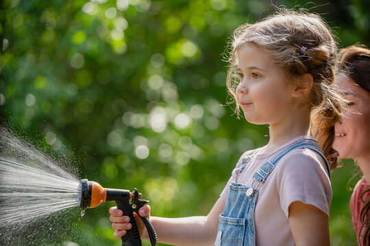 Young mother showing her little daughter how to water green lawn with garden hose.