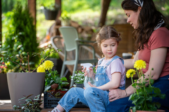 Young mother teaching her little daughter how to spray flowers on wooden terrace