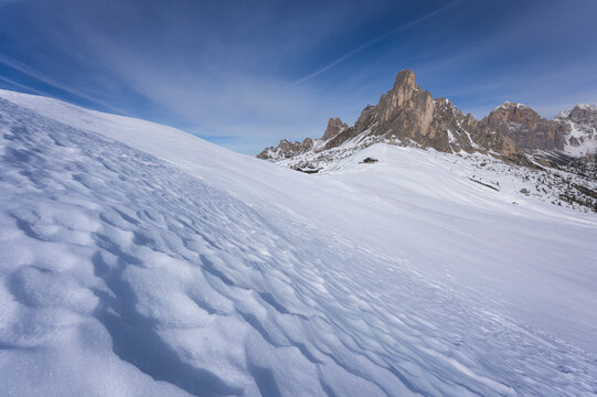 View of a pristine, wavy snowscape contrasting with sharp, rocky peaks under a brilliant blue sky, a remote hut visible, Belluno, Veneto, Italy.