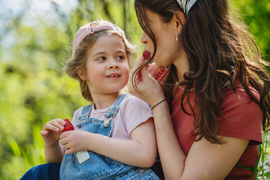 Happy mother and daughter eating fresh strawberries while sitting outdoor in summer garden