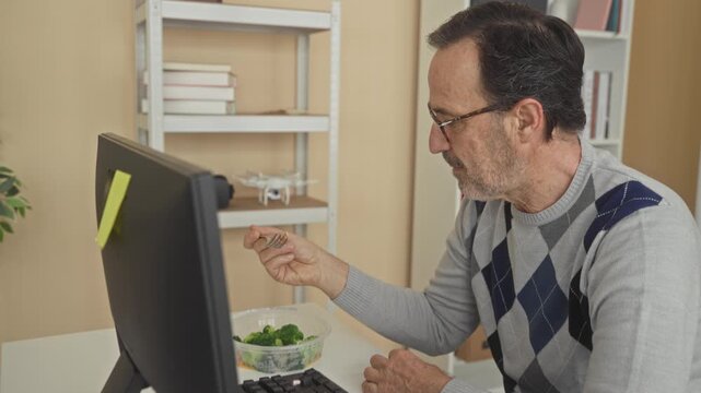 Man eating salad with fork, holding clear plastic container beside keyboard at computer desk in building; quiet concentration.