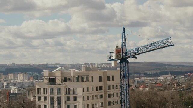 Large blue tower crane working on a construction site, rotating its jib over an urban landscape with apartment buildings against a background of cloudy sky, symbolizing urban development. timelapse