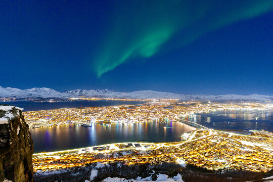 View of a breathtaking winter night over a sprawling city, its golden lights shimmering on the dark water beneath mesmerizing green Northern Lights, Tromsdalen, Troms, Norway.