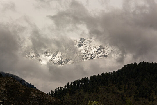 View of snow-capped peaks pierce through the heavy, low-hanging clouds, contrasting with the dark, forested hillsides below, Leepa, Azad Kashmir, Pakistan.