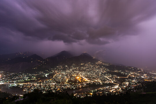 View of city lights twinkling beneath ominous, swirling clouds as lightning illuminates the mountains, a dramatic display of nature's power, Muzaffarabad, Pakistan.