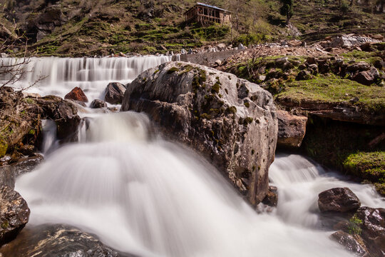 View of cascading white water rushes over jagged rocks, contrasting with the lush green hills and rustic cabin above the falls, Neelum Valley, Pakistan.