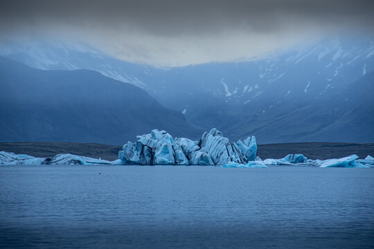 View of a blue iceberg floats serenely in the glacial lagoon against a backdrop of misty mountains, Glacier Lagoon, Sveitarfelagid Hornafjordur, Iceland.
