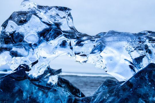 View of a glistening, translucent iceberg archway framing the distant, moody ocean horizon under a pale sky, Jokulsarlon, Sveitarfelagid Hornafjordur, Iceland.