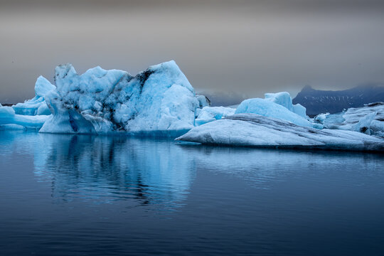 View of icebergs reflecting on the calm, dark waters under a muted sky, showcasing the serene yet stark beauty of nature, Glacier Lagoon, Iceland.