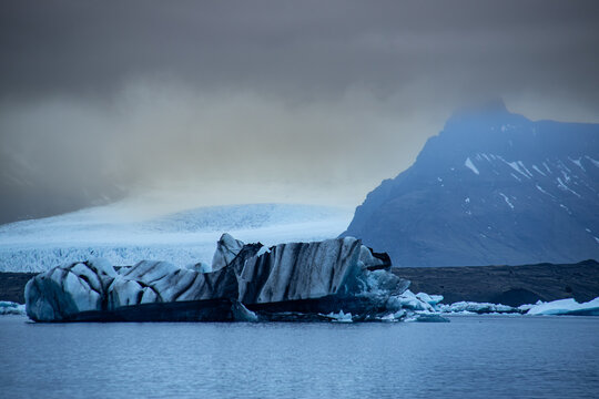 View of a striated ice block floating serenely on the glacial waters beneath a brooding sky, with mountains looming in the distance, Glacier Lagoon, Sveitarfelagid Hornafjordur, Iceland.